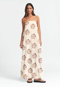 Häbemeeta maksikleit kreemivärvi kangast, millel on pruunid lillemustrid, lahtise lõikega ja küljeavad, koos mustade lamedate sandaalidega.