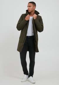Abrigo acolchado en verde oliva con un cuello alto, jeans negros y zapatillas blancas. El abrigo cuenta con un cierre de cremallera y botón.