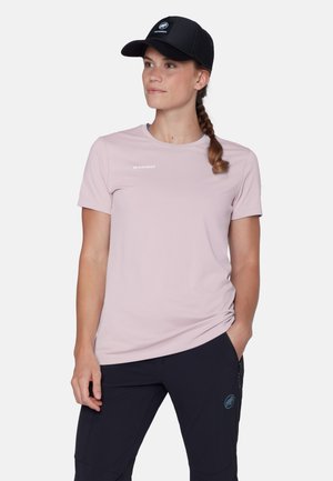 Frau trägt ein hellrosa Mammut-T-Shirt, eine schwarze Mammut-Kappe und dunkle Hosen, hat eine Hand in der Tasche und schaut zur Seite.