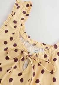 Gelbes ärmelloses Oberteil mit braunen, unregelmäßigen Punkten, gerüschtem Ausschnitt und gebundenem Schleifendetail an der vorderen Schlitzöffnung.