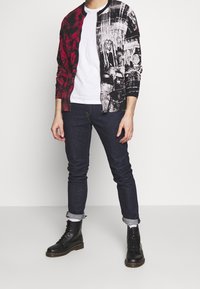 Red and black graphic cardigan with long sleeves, paired with a white T-shirt and dark jeans. Black lace-up boots complete the outfit.