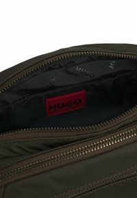 Interno di una borsa di tessuto marrone con etichetta rossa "HUGO". Dotata di chiusura a zip e texture liscia con una fodera a motivi sottili.
