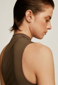 Top marrón sin mangas con un escote alto, fabricado con un tejido suave y con costuras de hombro visibles, llevado por una persona con cabello corto.