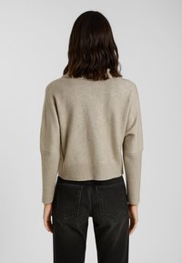 Donna con capelli mossi e scuri, che indossa un maglione beige a maniche lunghe e jeans neri a vita alta, in piedi di spalle contro uno sfondo semplice.