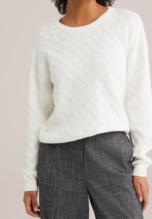 Femme portant un pull en maille blanche texturée avec un motif en losange et un pantalon à carreaux gris taille haute, torse visible.