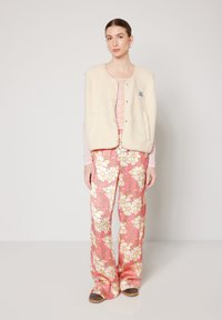 Fleece vest in crèmewit met knoopsluitingen, gecombineerd met roze bloemenbroek met grote witte madeliefjes en bruine schoenen.