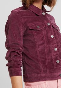 Cordjacke in tiefem Burgunderrot mit Brusttaschen, silbernen Knöpfen und umgeschlagenen Ärmeln. Weiche Textur mit strukturierter Passform.