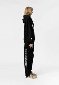 Sudadera con capucha negra y gran gráfico blanco, combinada con pantalones de chándal negros a juego que cuentan con estampado lateral y bolsillos. Zapatillas de color claro.