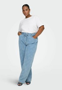 Femme les mains dans les poches portant une chemise blanche à manches courtes, un jean large bleu clair à motifs floraux et des talons ouverts bleu foncé.