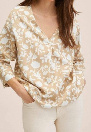 Kobieta w beżowej bluzce z dekoltem w serek, ozdobionej białym kwiatowym wzorem, oraz w jasnych spodniach, z rękami swobodnie w kieszeniach, na neutralnym tle.