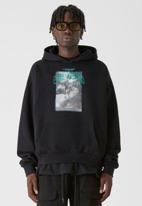 Melna kapuce ar priekšējo kabatu, grafiskais prints ar tekstu "Zaudētā Jaunatne" tirkīza krāsā, mīksta auduma, ribota apkakle un apakša. Slāņots izskats apakšā.