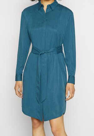 Vrouw draagt een teal kleurige overhemdjurk met lange mouwen, een kraag en een zelfbindende tailleceintuur.