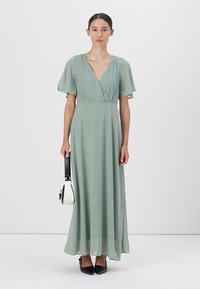 Lång, svävande klänning i mjuk grön textil, med fjäderlätta ärmar och V-ringning. Tillsammans med en liten handväska i svart och vitt.