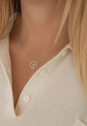 Silberne Halskette mit einem Baumdesign in einem runden Anhänger, platziert auf einem weichen beigen Pullover, mit sichtbaren Knopfdetails.