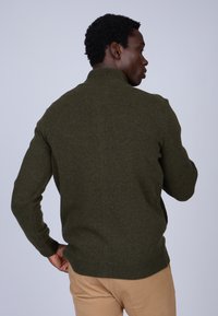 Pull olive en laine avec un col montant, texture en tricot côtelé et silhouette ajustée. Porté avec un pantalon beige, mettant en avant un style décontracté.