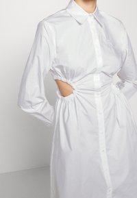 Vit skjortklänning med långa ärmar, elastiska sidokuttag och rynkade detaljer vid midjan. Slätt tyg med klassisk krage.