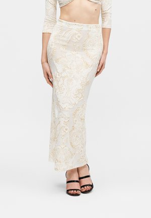 Moteris dėvi ilgą baltos ir aukso spalvų raštuotą sijoną, baltą trumpą viršutinę dalį ir juodus aukštakulnius sandalus, stovinti priešais vientisą foną.