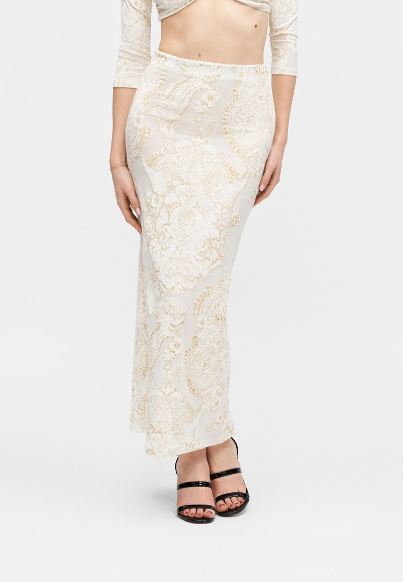 Γυναίκα που φοράει μακριά φούστα με λευκό και χρυσό μοτίβο, λευκό crop top και μαύρα ψηλοτάκουνα σανδάλια, στέκεται μπροστά σε απλό φόντο.