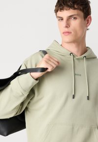 Sudadera verde claro con un bolsillo frontal y un sutil logo "BOSS". Modelo sosteniendo una bolsa negra, mostrando la tela y el diseño de la sudadera.