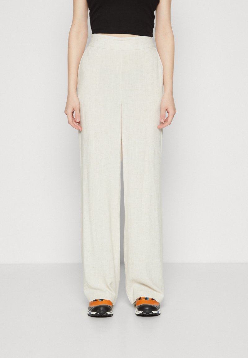 Femme portant un pantalon beige taille haute à jambes larges avec des plis, associé à un haut court noir et des baskets orange et noires.