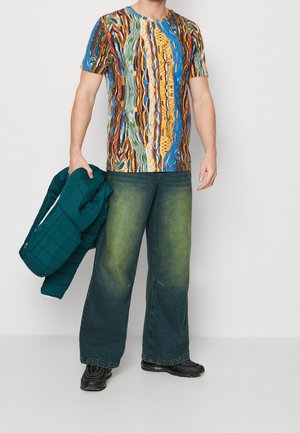 Mand med en multicolor mønstret kortærmet skjorte, grønne brede bukser, sorte sneakers, som holder en mørkegrøn dunjakke.