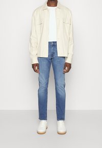 Světle béžová košile na knoflíky, bílý tričko, modré džíny a bílé tenisky se světlohnědou podrážkou. Košile má dvě přední kapsy.