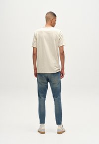 Beige pamut póló kerek nyakkivágással, világoskék farmernadrággal viselve. A farmernadrág enyhén szűkített fazonú, minimális tellálhatósággal.