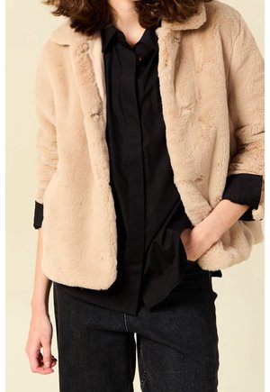Persoon draagt een beige jas van nepbont over een zwart gestoken overhemd en zwarte broek, één hand in de zak, staand tegen een neutrale achtergrond.