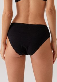 Zwarte katoenen slip met geschulpte kantafwerking langs de tailleband. Zachte textuur, volledige dekking, naadloze achterkant en minimale stiksels.