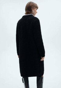 Cappotto teddy nero, lungo fino al ginocchio, tessuto strutturato, maniche lunghe, tasche laterali, colletto con dettaglio interno grigio, superficie liscia.