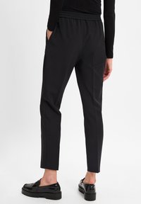 Persoon in zwarte, op maat gemaakte enkellange broeken en glanzende zwarte loafers, staand met één hand in de zak, op een witte achtergrond.