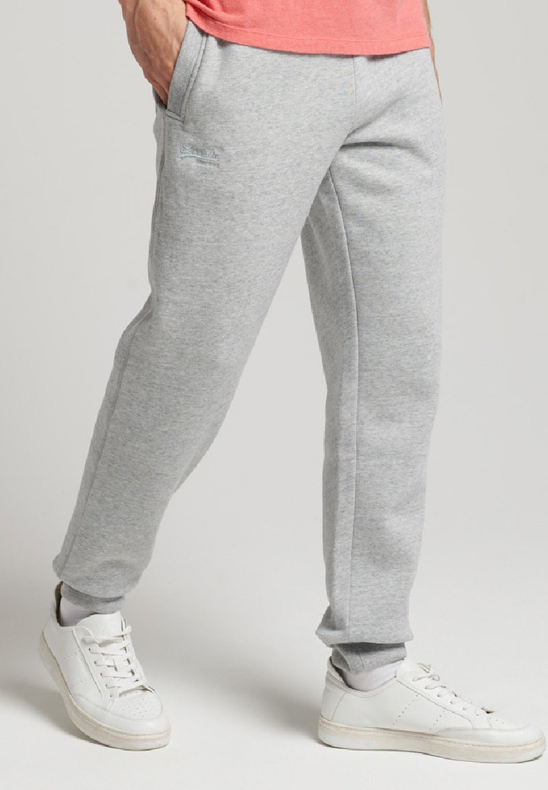 Pantaloni della tuta grigi realizzati in materiale morbido, caratterizzati da un design affusolato, tasche laterali e polsini a costine alla caviglia. Abbinati a scarpe da ginnastica bianche.