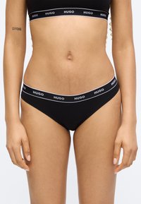 Γυναικείο σώμα με μαύρα εσώρουχα και αθλητικό σουτιέν με το λογότυπο Hugo, με τατουάζ "DIVINE" στο αριστερό αντιβράχιο εμφανές.