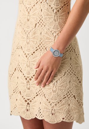 Orologio da polso blu con quadrante rotondo luccicante e cinturino blu abbinato, posizionato su un Abito in crochet beige con motivi di farfalle.