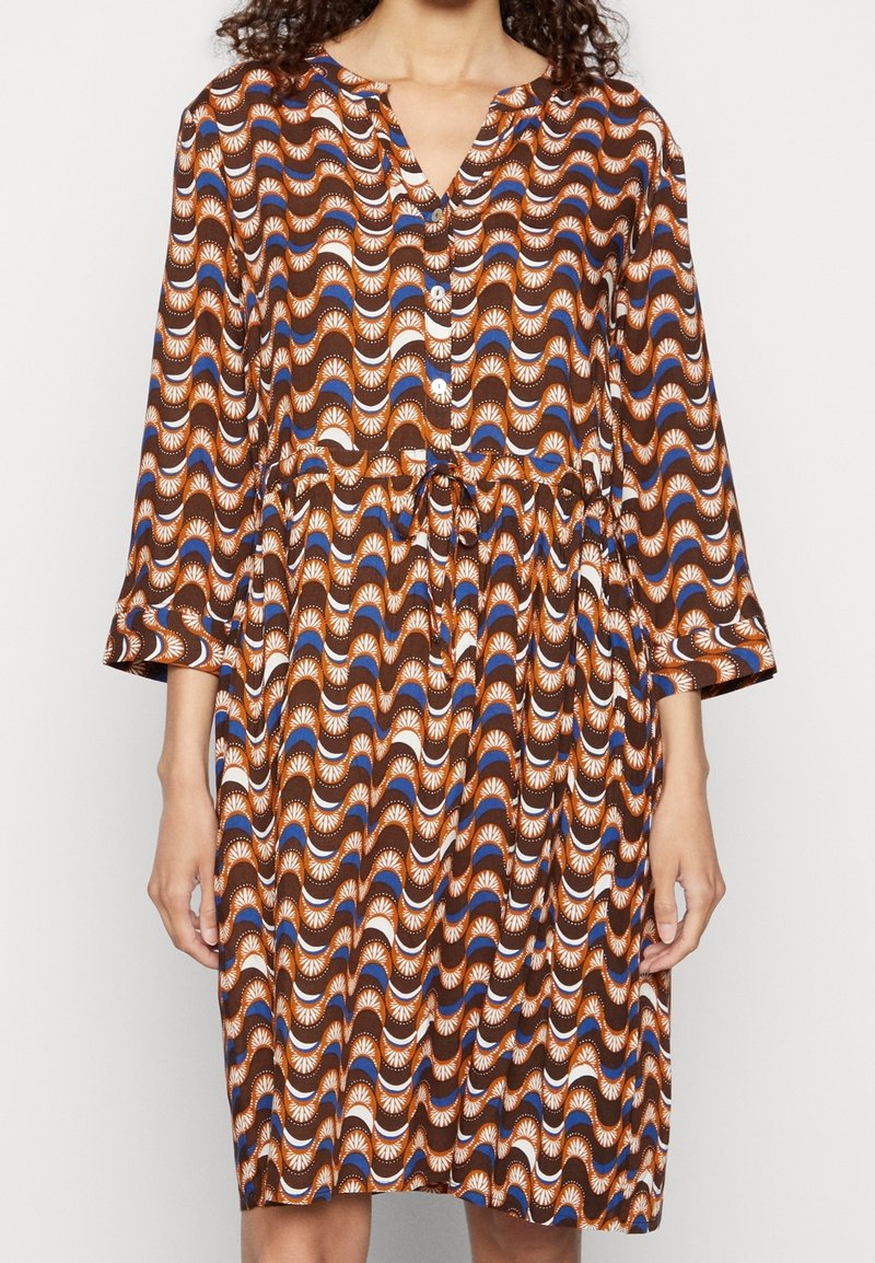 Femme portant une robe à motifs marron, bleu et blanc, longueur genou, avec des manches trois-quarts et des boutons sur le devant.