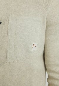 Tissu tricoté beige avec une poche avant portant une petite étiquette blanche brodée d'une lettre A stylisée marron.