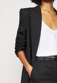 Blazer nero su misura con maniche arrotolate, indossato sopra una maglietta bianca a V. Cintura nera con fibbia rotonda dorata, tessuto strutturato. Design semplice.