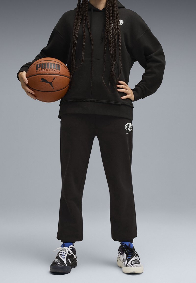 Personne portant un sweat à capuche noir et un pantalon de jogging tient un ballon de basket Puma d'une main, l'autre main sur la hanche.