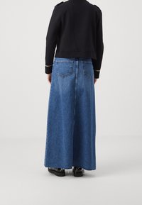 Personne portant une veste noire et une longue jupe en jean bleue avec des poches à l'arrière, debout devant un fond clair uni.