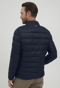 Marineblå puffjakke med horisontalt quiltet design, høy krage og en liten trikolor aksent på baksiden, laget av glinsende, lett materiale.