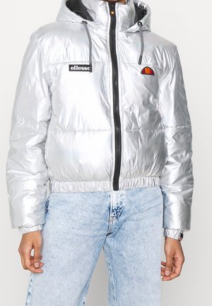 Person wearing a silver puffer jacket with a hood, black zipper, and ellesse logos on chest and denim jeans.