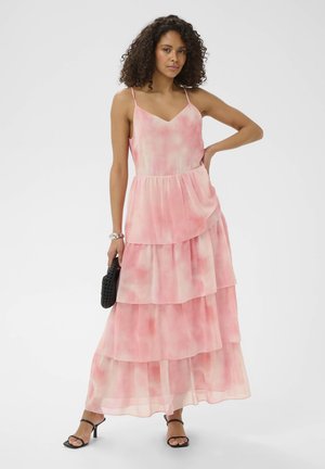 Femme aux cheveux bouclés portant une robe maxi rose à volants sans manches et des sandales à talons noires, tenant une pochette noire tissée.