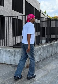 Weißes Kurzarmhemd, blaue weite Jeans und schwarze Schuhe. Ein pinkfarbener Bandana auf dem Kopf. Beton-Hintergrund mit geometrischen Designs.