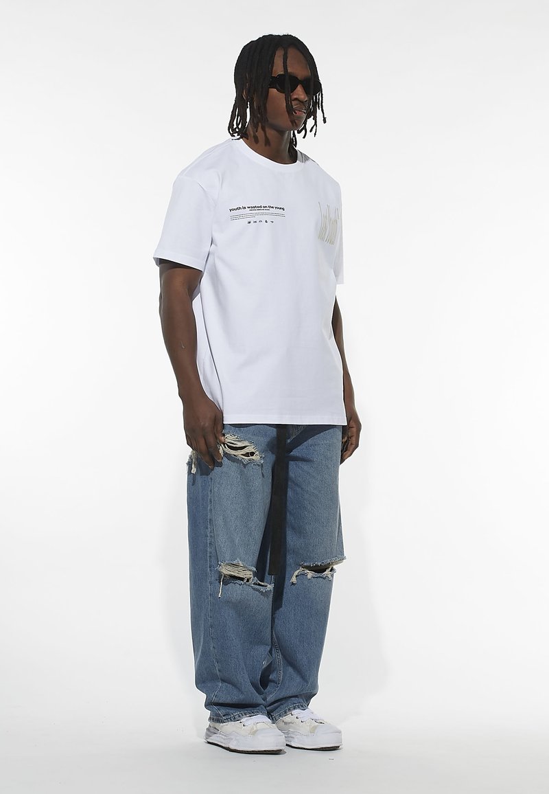 White t-shirt with text and graphic detail on the chest, paired with distressed blue jeans and white sneakers. Model wearing sunglasses.