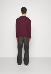 Bordeauxroter Pullover mit langen Ärmeln, Rundhalsausschnitt und gerippten Bündchen; kombiniert mit dunkelgrauen Jeans und weißen Turnschuhen; Rückansicht.
