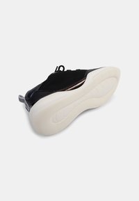 Chaussure de sport noire avec une tige en maille, semelle en caoutchouc blanche et un motif texturé. Dispose d'une languette à l'arrière pour un enfilage facile.