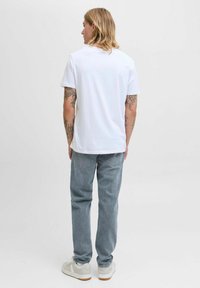 Camiseta blanca de algodón de manga corta, combinada con jeans de mezclilla azul claro y zapatillas blancas, con tatuajes visibles en los brazos.