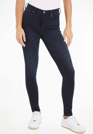 Vaqueros ajustados de color azul oscuro con una cintura alta, hechos de mezclilla. Las características incluyen un cierre de botón, presillas para cinturón y bolsillos delanteros. Combinados con zapatillas blancas.