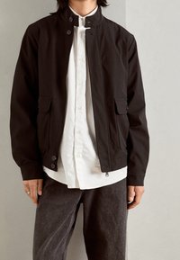 Person som bär en svart zip-up-jacka över en vit skjorta med knappar och mörkgrå jeans, stående mot en ljusgrå bakgrund.