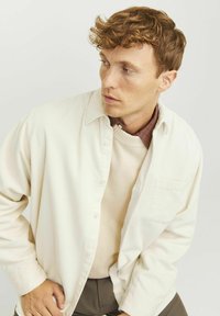 Camicia di corduroy bianca sovrapposta a un maglione beige, con colletto a scacchi. Il modello indossa pantaloni scuri e ha i capelli mossi.